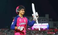 Cricketer in a pink jersey and helmet raises a bat in celebration on the cricket field.