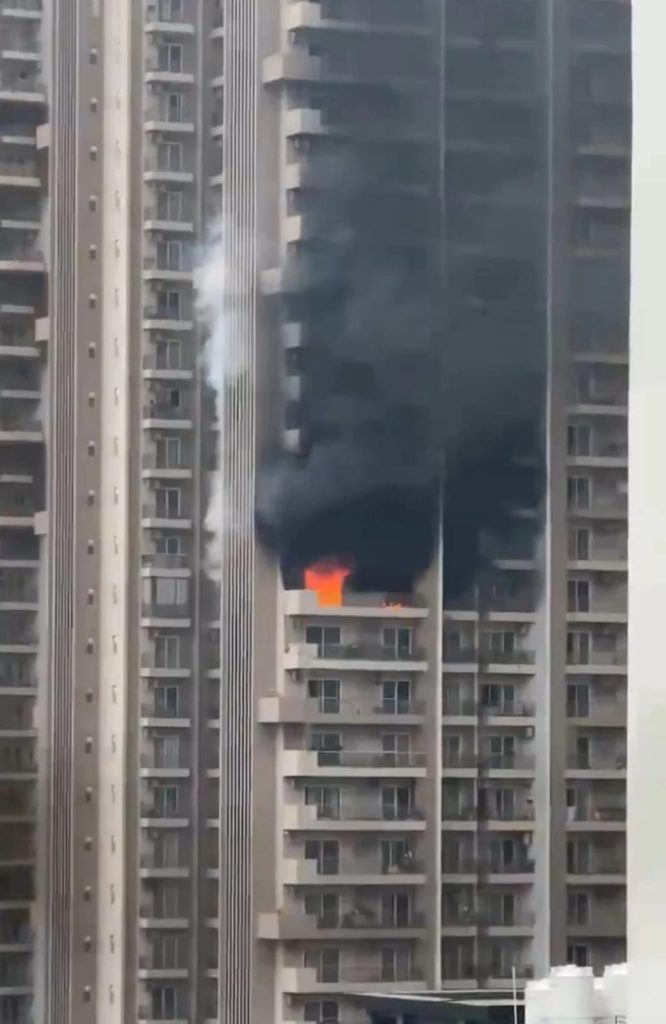 High-rise apartment building releasing thick black smoke with bright orange flame at a balcony area on one floor.