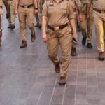 Group of police officers in khaki uniforms walking down a paved sidewalk together in an urban setting.