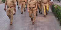 Group of police officers in khaki uniforms walking down a paved sidewalk together in an urban setting.