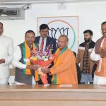 A group of men in formal and traditional attire present a flower bouquet to a Buddhist monk in orange robes during a BJP event, with a party symbol on the wall behind them.