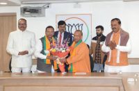 A group of men in formal and traditional attire present a flower bouquet to a Buddhist monk in orange robes during a BJP event, with a party symbol on the wall behind them.