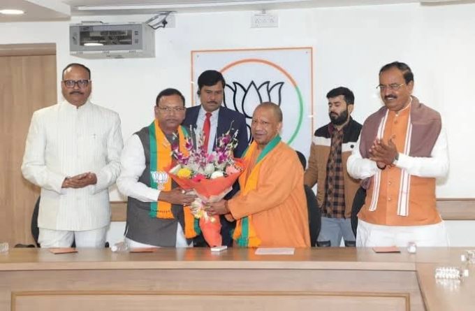 A group of men in formal and traditional attire present a flower bouquet to a Buddhist monk in orange robes during a BJP event, with a party symbol on the wall behind them.