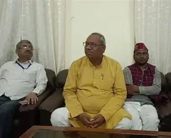 Three men sit on a brown sofa in a room with white curtains; center man wears a yellow kurta, others in white shirt and red cap.