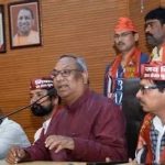 Man in a maroon shirt speaks into a microphone at a press conference, flanked by supporters wearing saffron scarves; portraits hang on a wooden wall behind them.