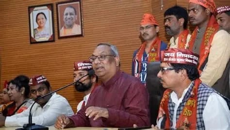 Man in a maroon shirt speaks into a microphone at a press conference, flanked by supporters wearing saffron scarves; portraits hang on a wooden wall behind them.