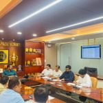 Professionals seated around a long conference table in a wood-paneled office, reviewing documents with a display of awards on the shelf nearby and a wall-mounted screen in the background.