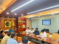 Professionals seated around a long conference table in a wood-paneled office, reviewing documents with a display of awards on the shelf nearby and a wall-mounted screen in the background.