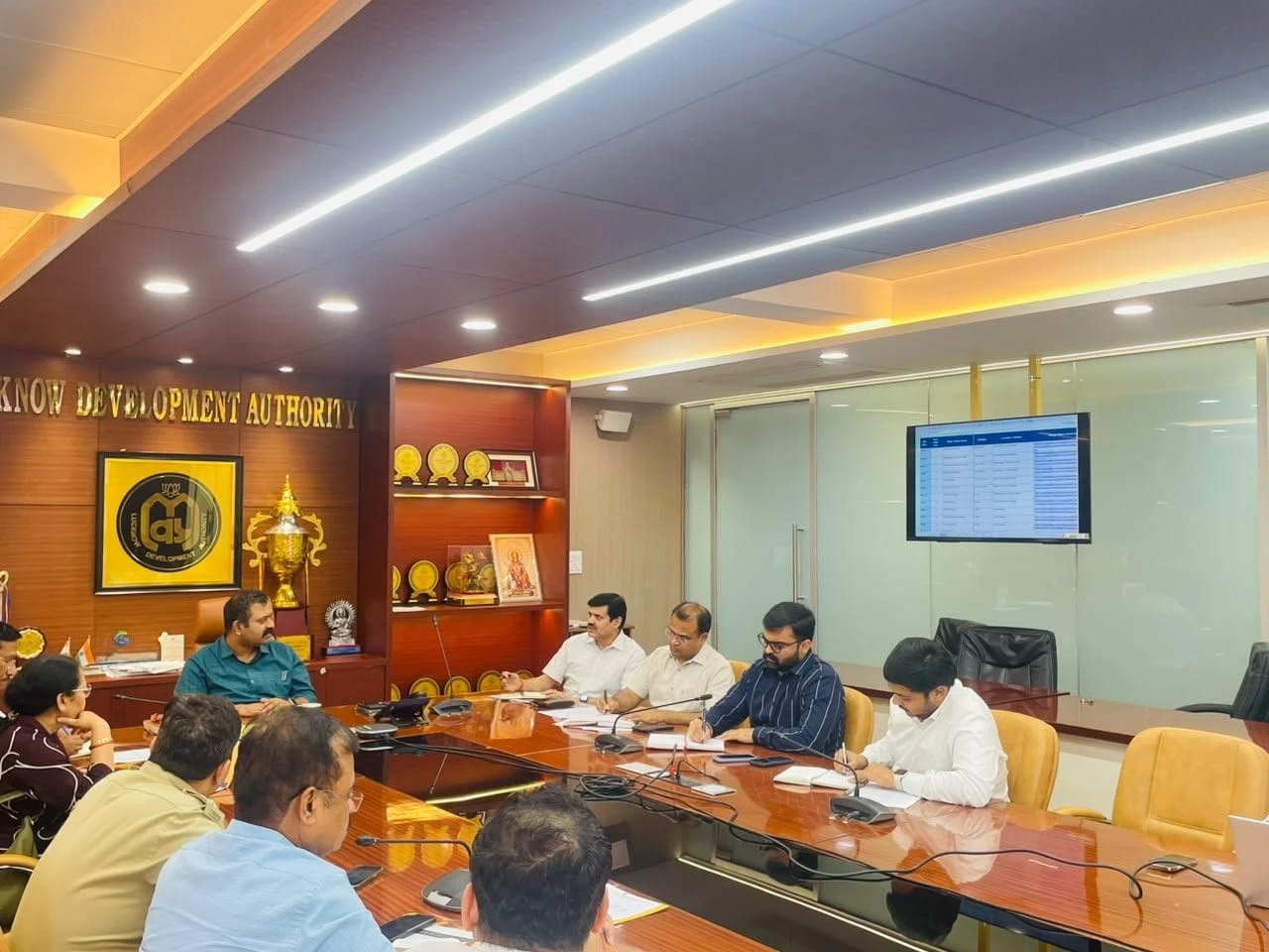 Professionals seated around a long conference table in a wood-paneled office, reviewing documents with a display of awards on the shelf nearby and a wall-mounted screen in the background.