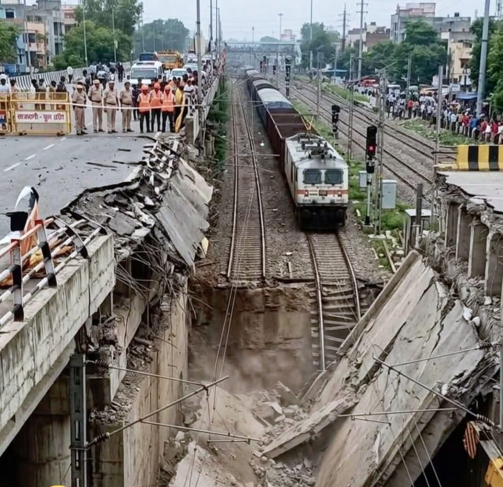 Train runs along a severely damaged railway bridge; collapsed concrete spans, dust rising, with officials and onlookers behind barricades on the road above.