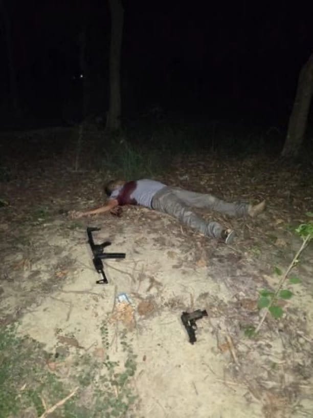 Man lying motionless on the forest floor at night, with several firearms scattered nearby (one rifle and a handgun).