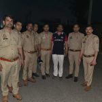 Group of eight men standing on a dimly lit street; seven are in khaki police uniforms, one man in a black and red Hackett polo shirt and white pants is in the center, posing for a photo at night.