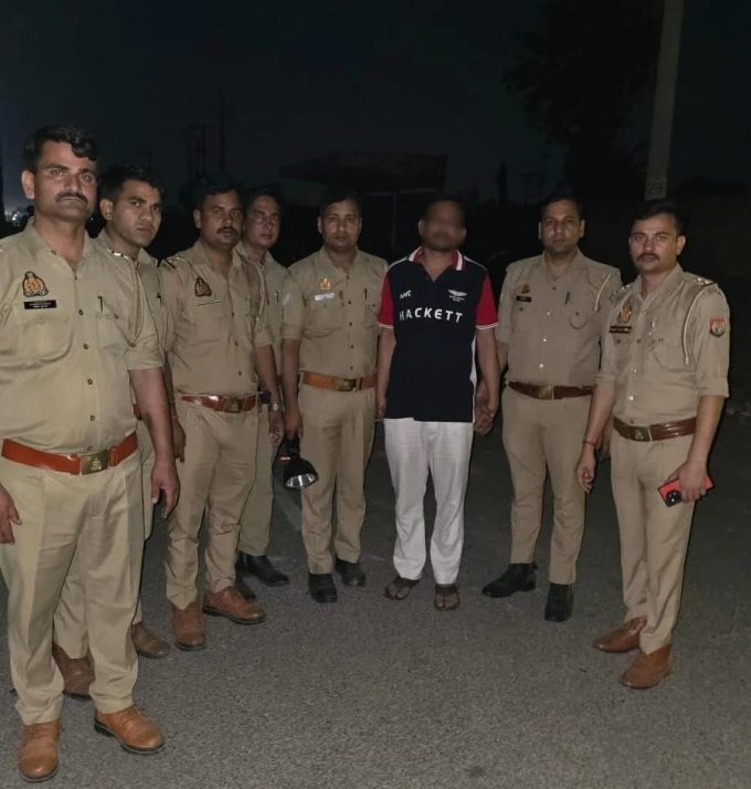 Group of eight men standing on a dimly lit street; seven are in khaki police uniforms, one man in a black and red Hackett polo shirt and white pants is in the center, posing for a photo at night.