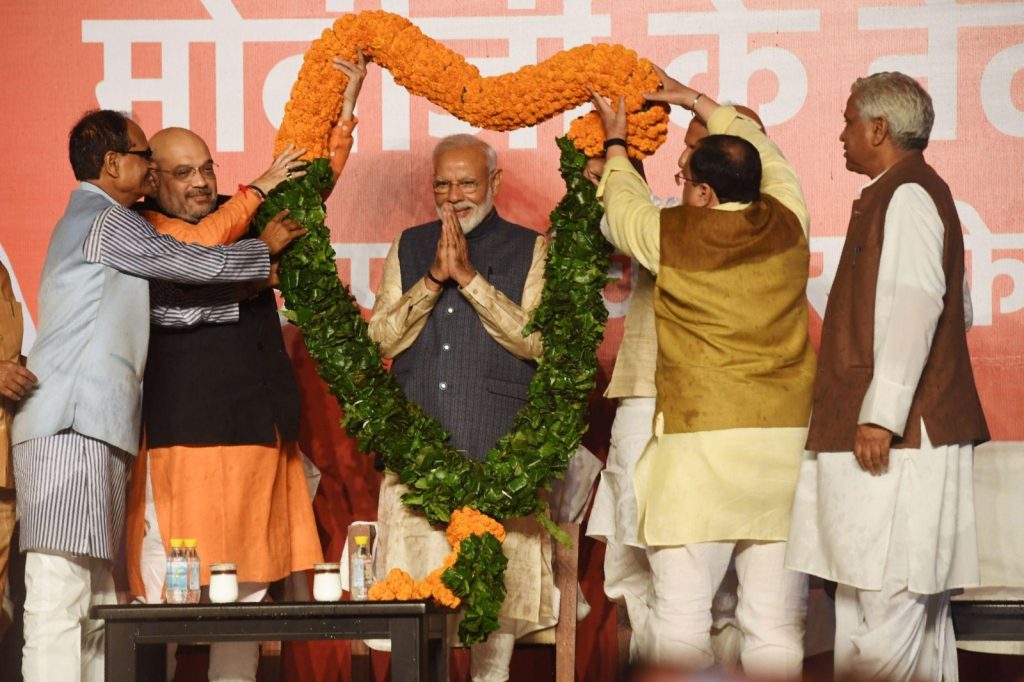 Man in a beige kurta and gray vest standing with hands pressed together as others drape a large orange flower garland around him at a ceremony.