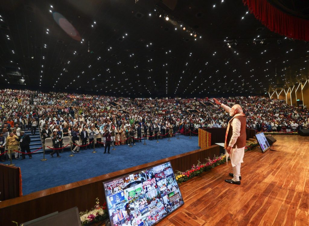 Speaker on stage addressing a large audience in a packed auditorium, with screens and bright overhead lights.