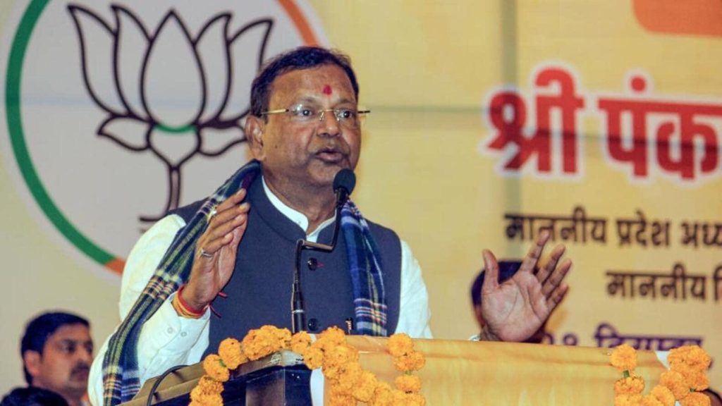 Indian man in a dark vest and white kurta speaks at a podium decorated with marigold garlands during a political event