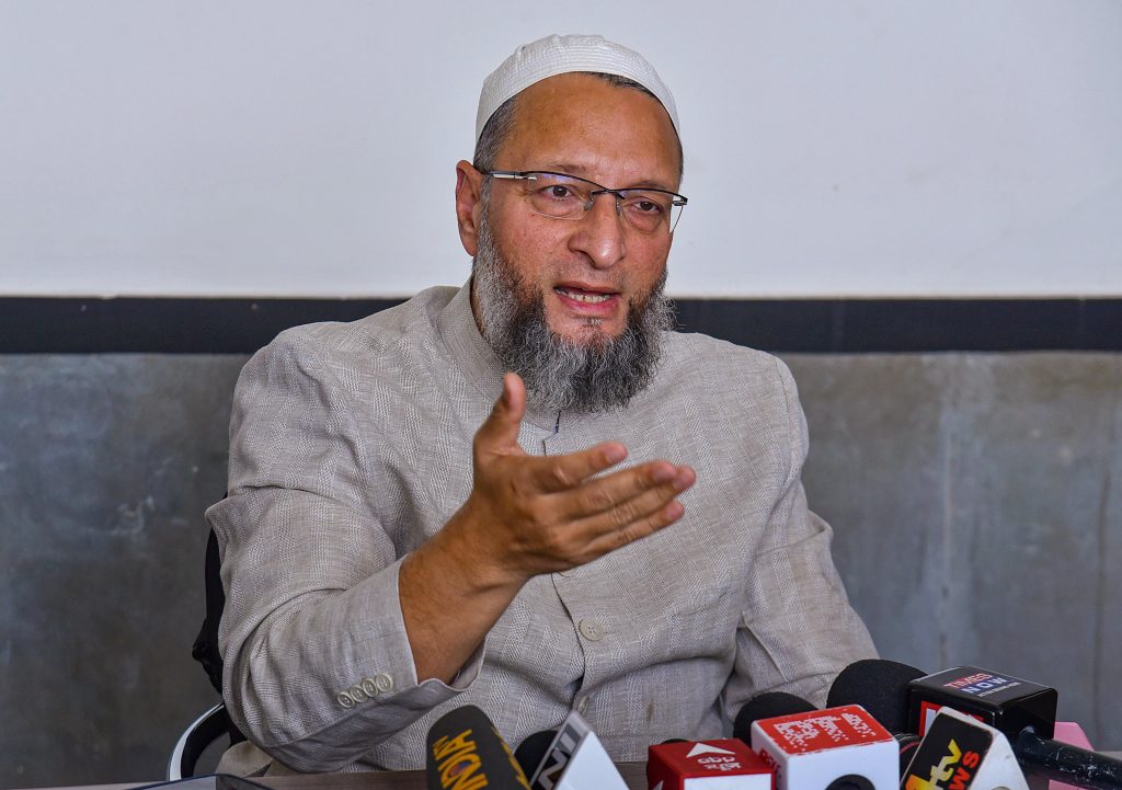 Bearded man in a light gray suit and white cap speaks at a press conference, gesturing with his hand amid multiple microphones on the table.