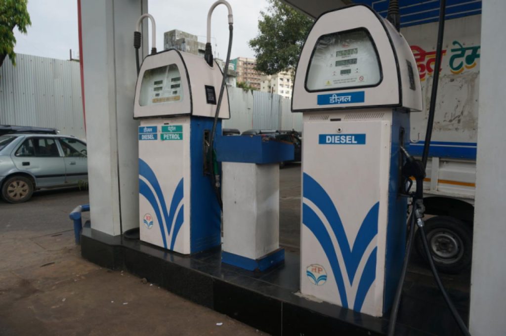 Diesel and petrol fuel pumps at a gas station with blue and white branding, a car in the background, and hoses hanging from the pumps, station canopy visible above.