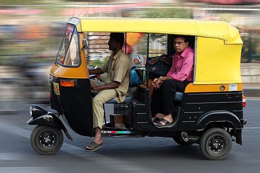 Auto rickshaw (tuk-tuk) in motion on a street; driver in a beige shirt in front and a passenger in a pink shirt seated in the back.