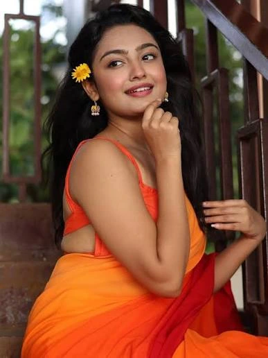 Young woman in an orange saree sitting on wooden stairs, with a yellow flower in her hair and a warm smile.