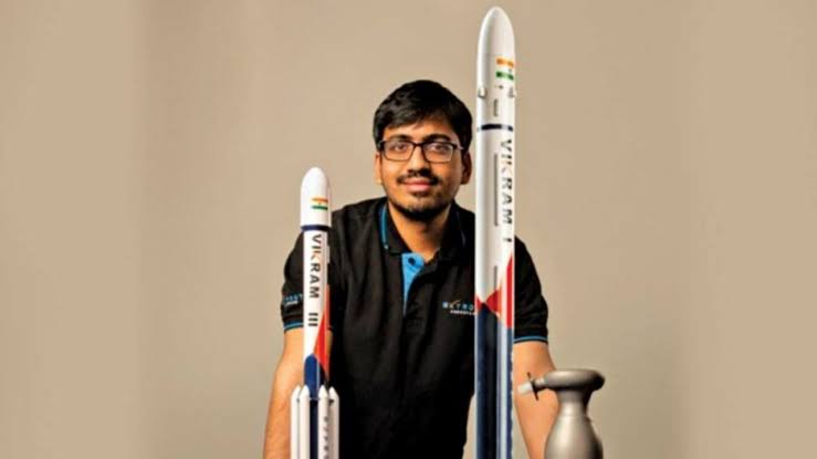 images 11 - The Journalist News Portrait of a man with glasses smiling, standing behind two tall model rockets on a neutral beige background, one rocket taller than the other.