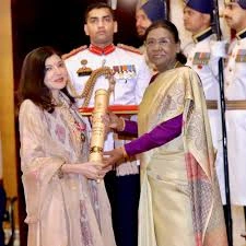 Woman in a pale pink saree hands a tall ceremonial torch to another woman in a lavender outfit on stage, with uniformed guards in the background.