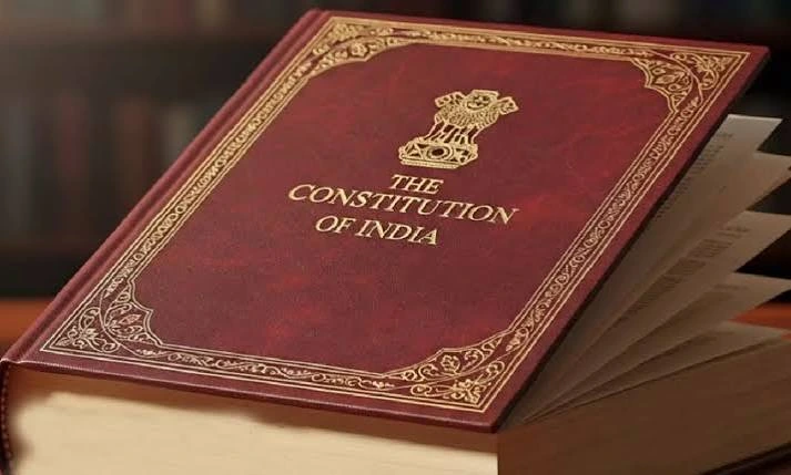 Maroon leather-bound copy of The Constitution of India with gold embossing, resting on an open book rack.