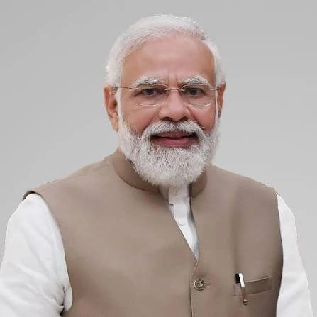 Older man with white beard and glasses, wearing a beige waistcoat over a white kurta, smiling at the camera.