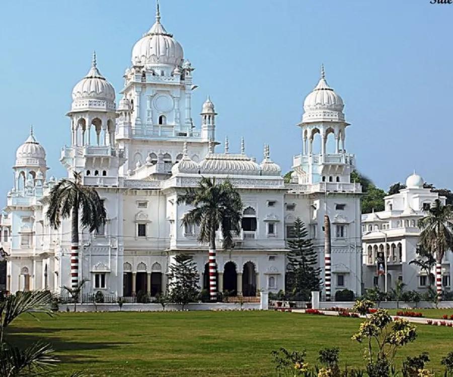 White ornate palace with multiple domes and arches, set in a well-kept lawn with palm trees under a blue sky