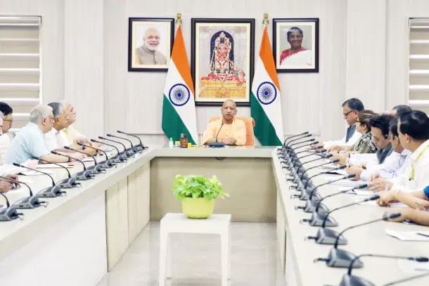 Central official at the head of a long conference table in a government meeting, with Indian flags and portraits on the wall behind.