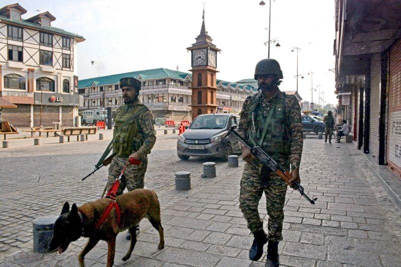 Two armed soldiers in camouflage walk a dog on a red harness through a town square, clock tower in the background.