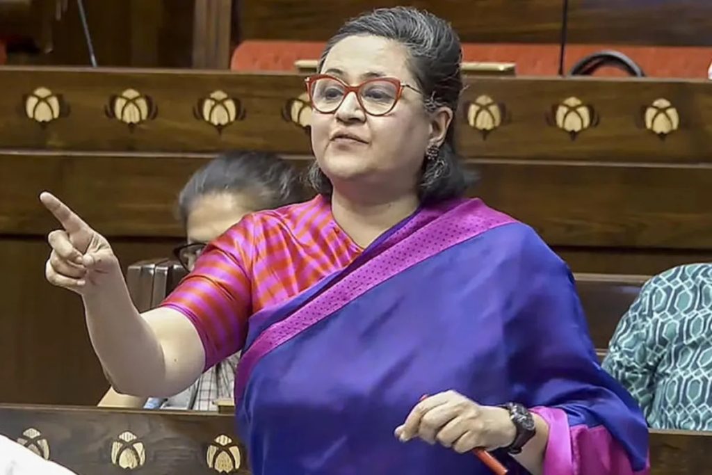 Woman in a purple sari with red glasses points her finger while speaking in a legislative chamber.