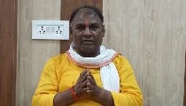 Man in a yellow kurta with a white scarf, standing indoors with hands folded in a Namaste greeting.