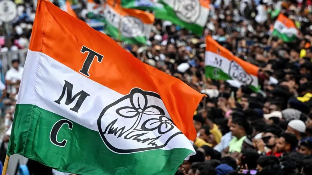 Flag of the Indian political party TMC waving above a crowded rally scene, saffron-white-green with a flower emblem at center-right.
