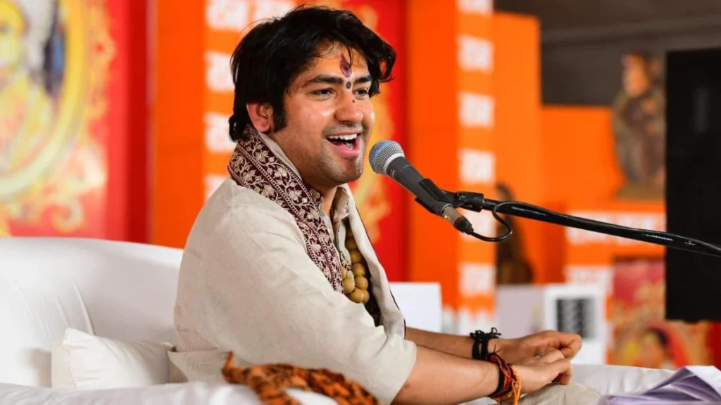 Smiling man in traditional beige outfit and bead necklace speaking into a microphone while seated on a white couch at a cultural event.