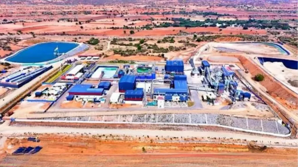 Aerial view of a blue-roofed industrial complex in a dry desert, with blue tanks, pipes, and fenced perimeter.