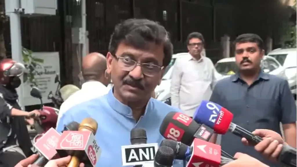 Man in a blue kurta speaks to multiple reporters holding microphones from various news channels.