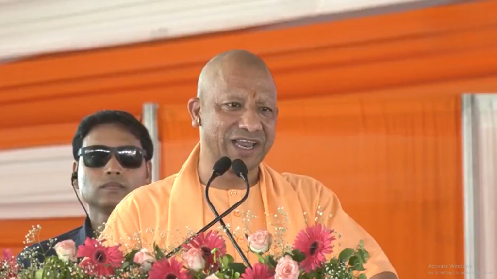 Bald man in a saffron robe speaks into two microphones at a podium, with a floral arrangement and an orange backdrop behind him.