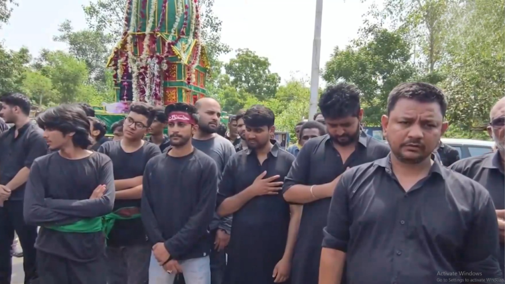 Group of men in black clothing standing in line during a funeral or religious procession, with a colorful decorated chariot in the background