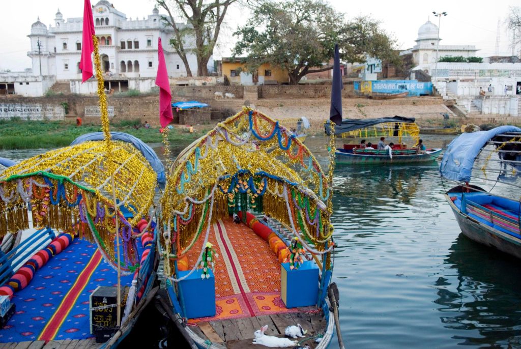 Colorfully decorated festival boats adorned with garlands on a calm river, with white temples and trees in the background.]','Colorfully decorated festival boats adorned with garlands on a calm river, with white temples and trees in the background.