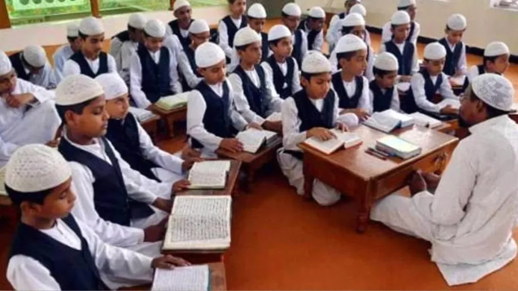 Class of young boys in white caps and navy vests reading from Qurans at desks as a teacher leads the session at the front of the room.
