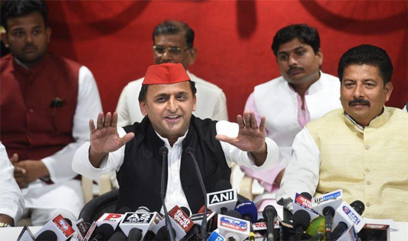 Man in a red cap speaking at a press conference, gesturing with both hands, with many microphones in front and others listening in the background.