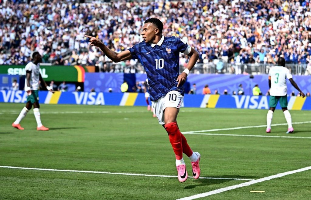 Soccer player in a blue jersey number 10 raises an arm and points as he celebrates on a green pitch, crowd in the stands behind him.