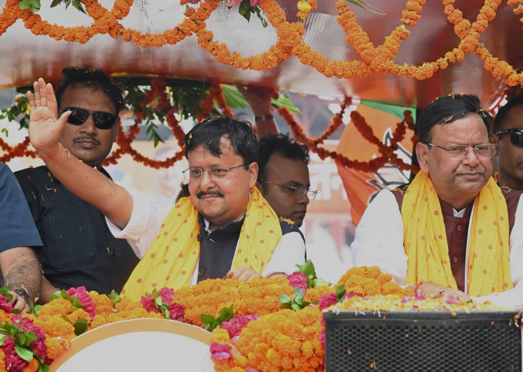 Group of men in a parade booth adorned with orange marigold garlands and yellow scarves, one man waving to the crowd.