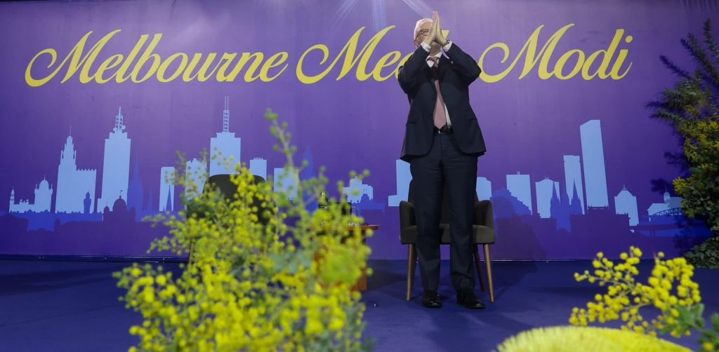 HMx_QRcbsAA2W9G - The Journalist News Man in a dark suit stands on a blue stage, clapping, with a purple backdrop that reads 'Melbourne Modi' and a city skyline silhouette behind him.