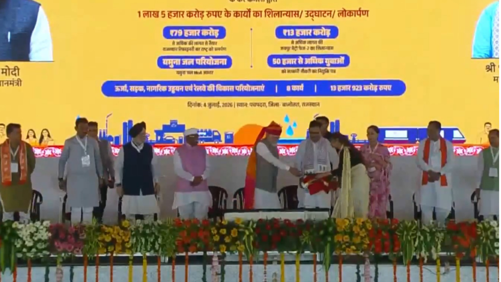 Dignitaries on a stage at a formal ceremony with a yellow backdrop and flower garlands, as a woman in saree presents an item to a man in traditional attire.