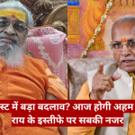 Two Indian men sit side by side in an ornate room; one with orange robes and a white beard, the other in a beige jacket gesturing toward the camera.