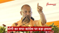 Man in orange robes speaks at a dais with two microphones, raising his index finger as he addresses the crowd; floral decor surrounds the podium, with a Hindi news banner at the bottom.