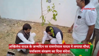 Three men plant a young sapling in a dusty outdoor area, one tending the sapling while another sits nearby; a green water container is visible, and a red Hindi news banner runs across the bottom.