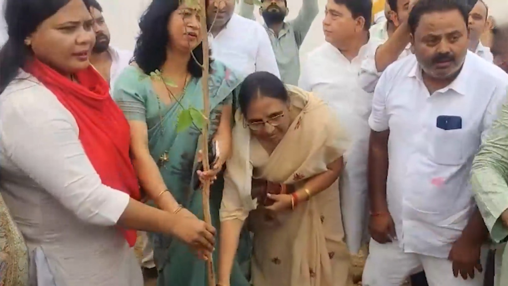 Group of people in traditional Indian attire around a sapling as a woman in a beige saree bends to touch the ground during a tree-planting ceremony.